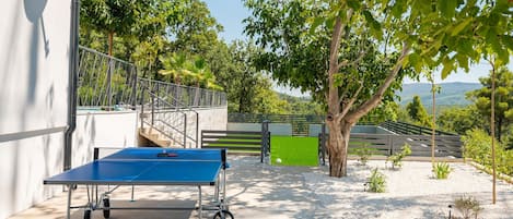 Mesa de ping pong al aire libre con baranda de metal y vista a un campo verde y árboles.