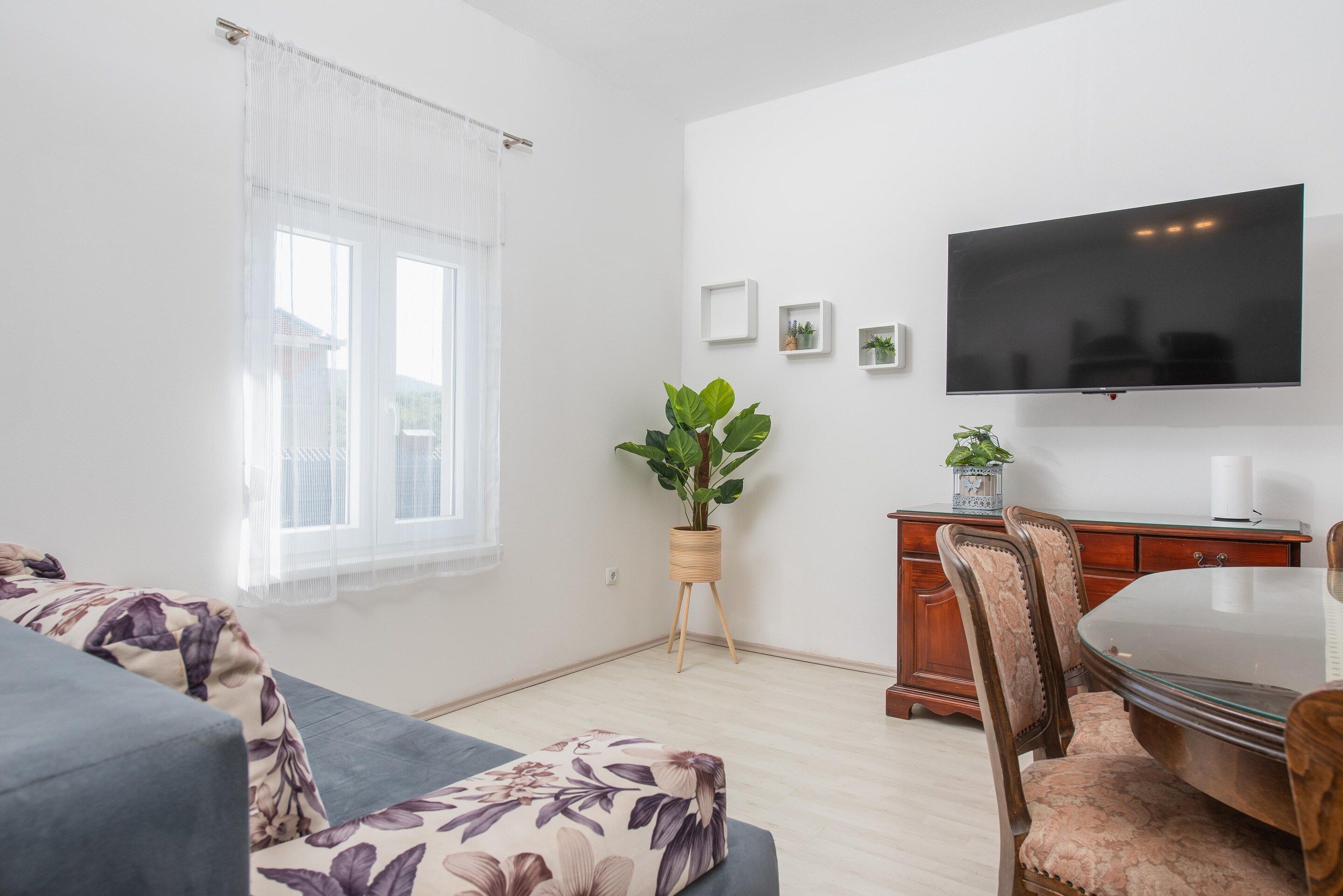 Un salon comprenant un canapé, une table à manger avec des chaises, un téléviseur fixé au mur et une fenêtre avec des rideaux.