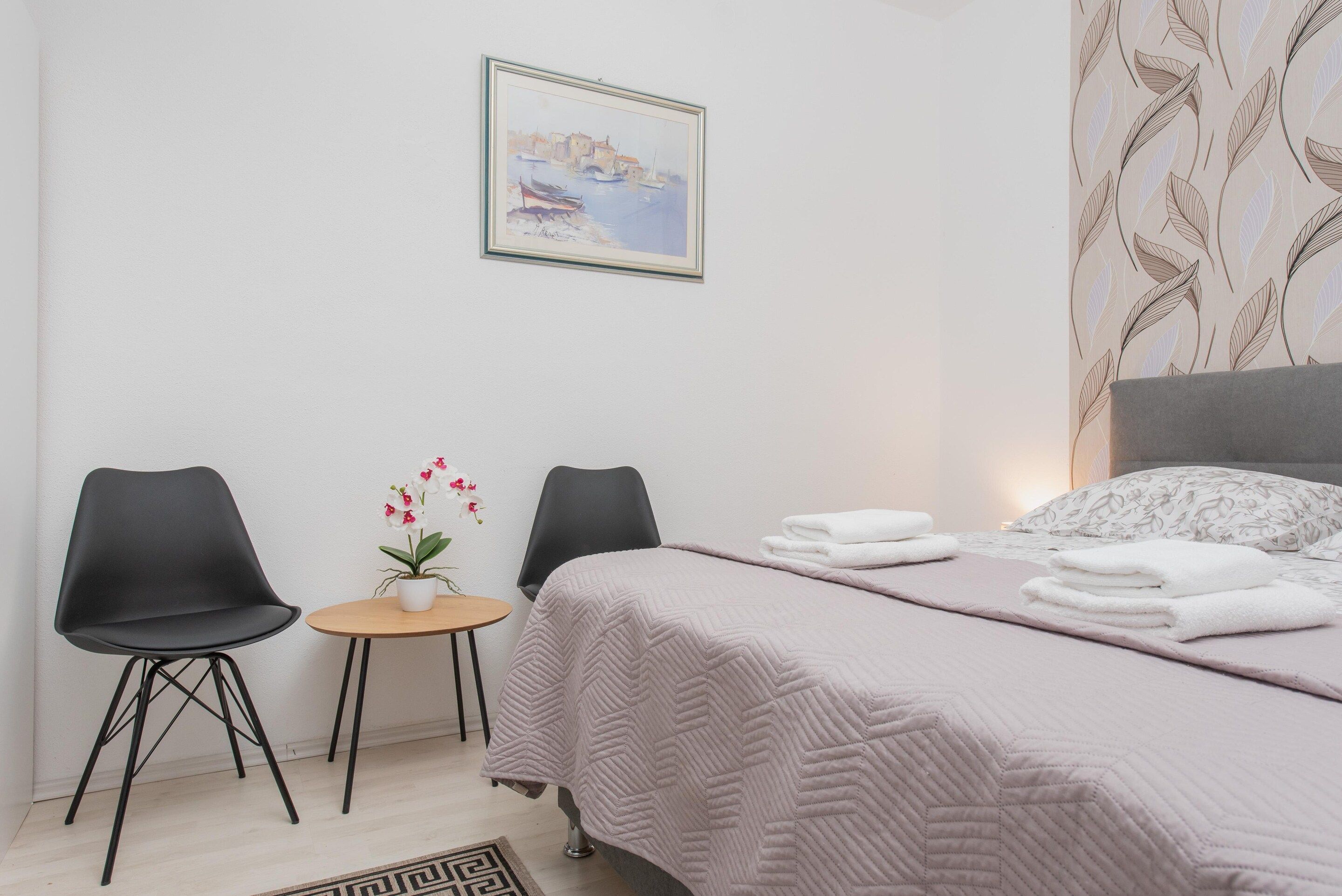 A hotel room with a bed, two chairs, a small table with a potted plant, and a painting on the wall.