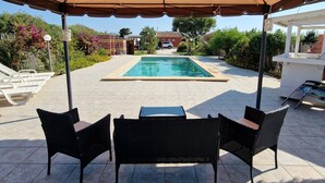 A covered outdoor seating area with two wicker chairs and a table, a pool in the background, and a tiled patio. - Vacation home with pool and within walking distance to the sea (Pachino)