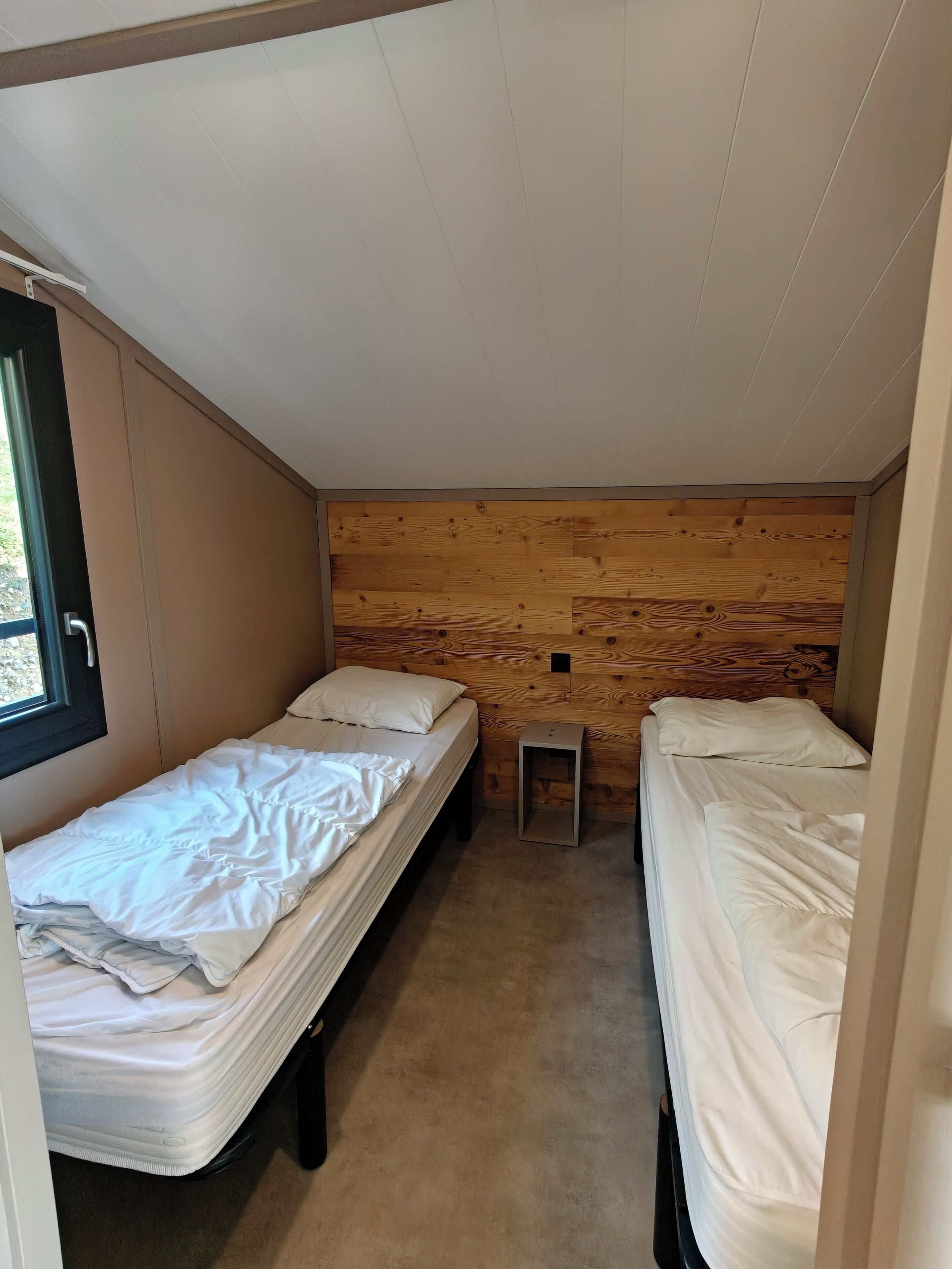 Une chambre de taille modeste, avec deux lits simples, une petite fenêtre et un mur en bois.