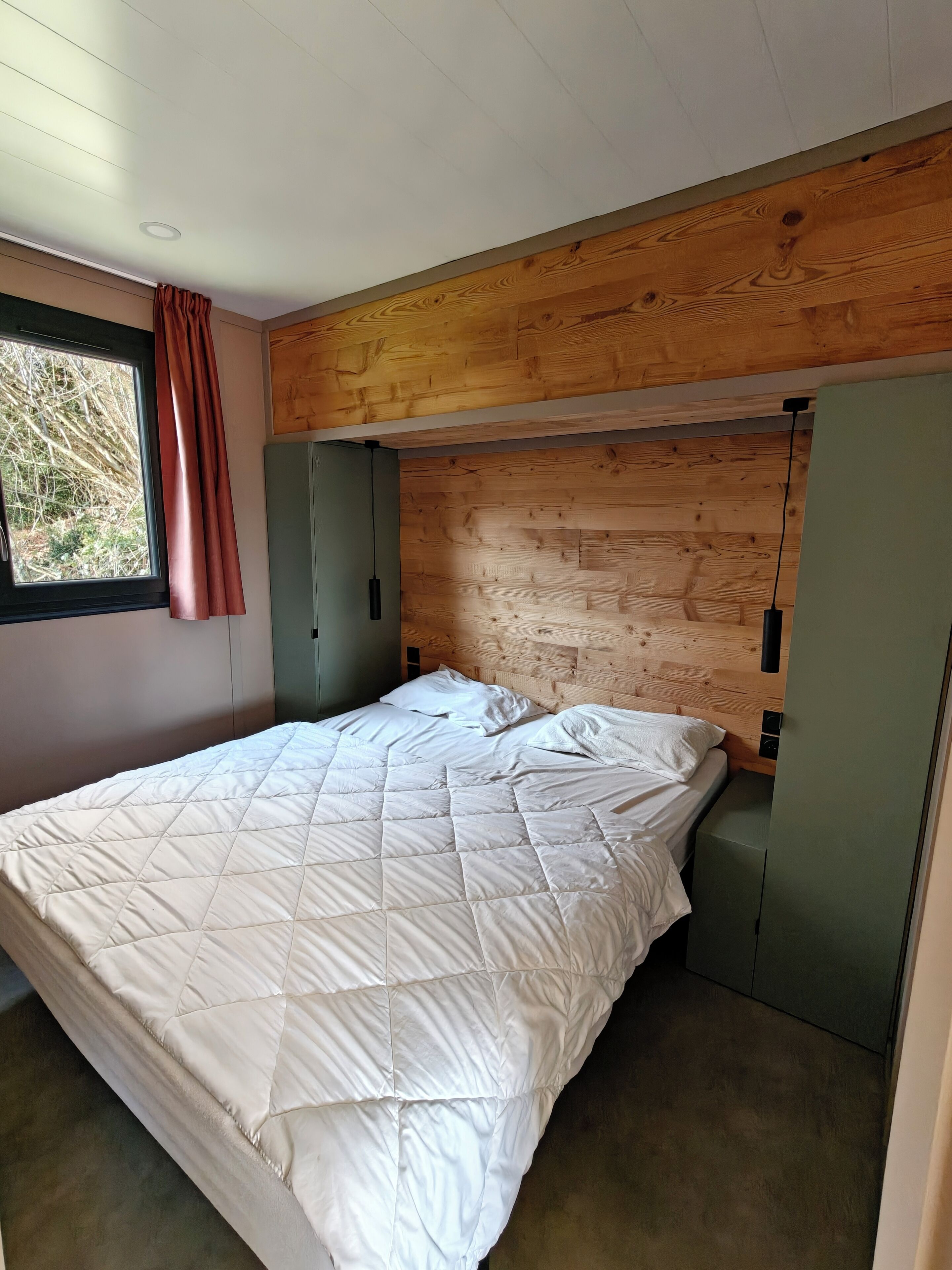 Ein Schlafzimmer mit einem hölzernen Kopfende, einem Bett mit weißer, gesteppter Bettdecke, einem Fenster mit Vorhang und einem grünen Kleiderschrank.
