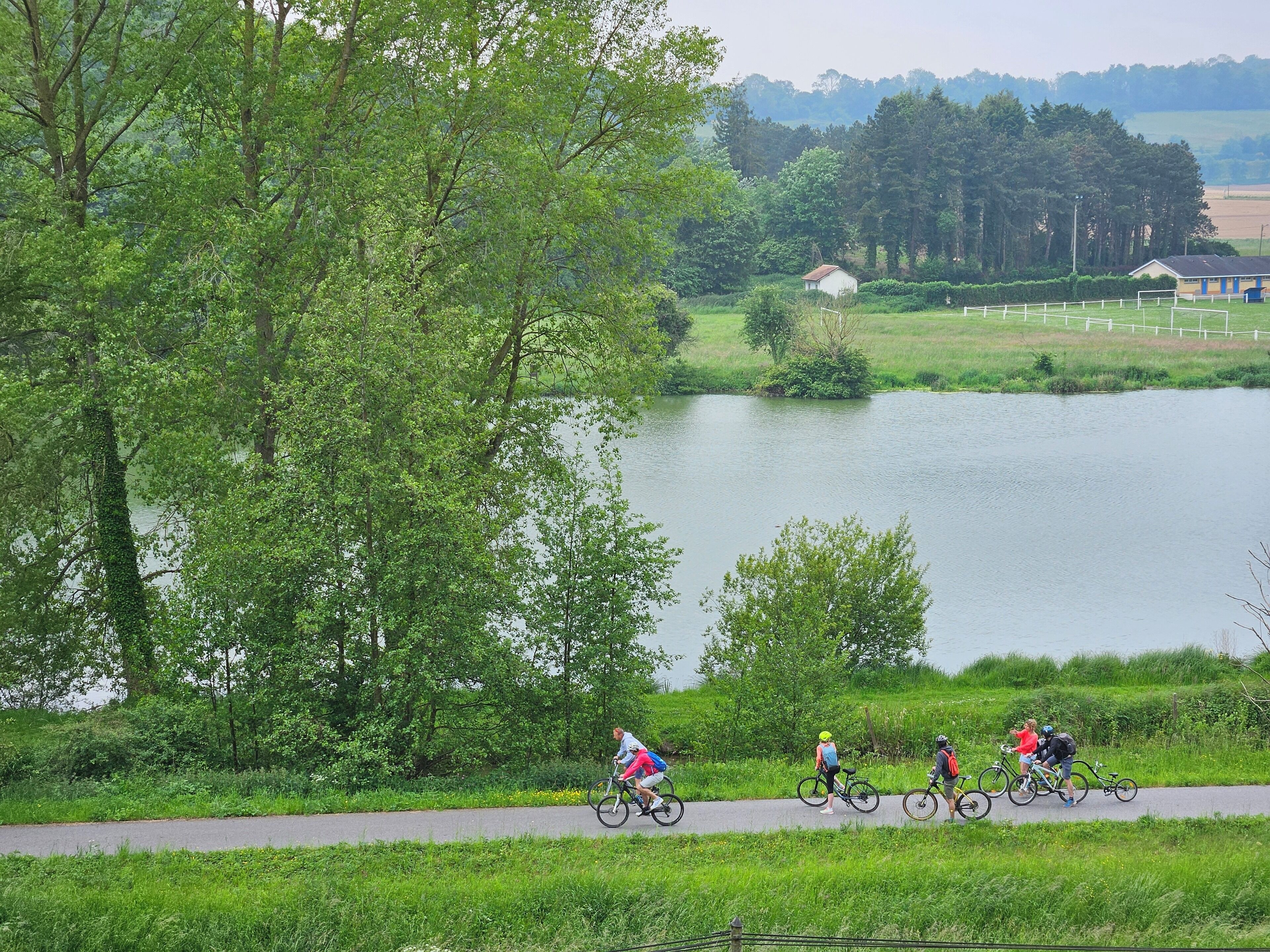 Une piste cyclable asphaltée longe un lac où l’on peut voir des cyclistes.