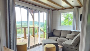 A spacious living area with a wooden floor, a grey sectional sofa, a small round ottoman, and a black coffee table. Large windows provide a view of the outdoors.