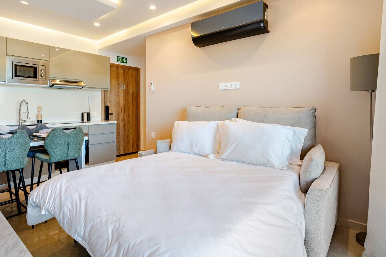 A hotel room with a large bed, a desk with chairs, a kitchenette, and a wall-mounted air conditioner.