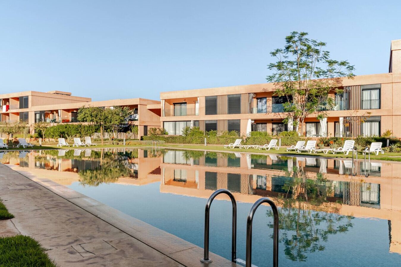 Un hôtel moderne doté d’une piscine, de chaises longues et dont l’image se reflète clairement dans l’eau.