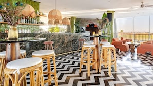 Eine Barbereich mit Schachbrettfußboden, Korbstühlen und einer Bar mit einer grünen Mosaikwand und Pfauenornamenten.