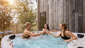 Trois femmes dans un jacuzzi, avec un décor en bois, entourées d’arbres et d’un mur de pierres.