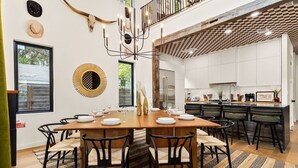 Un espace repas moderne avec une table en bois dressée pour six personnes, entourée de chaises noires. Un élément décoratif orne le mur au-dessus de la table, et un lustre est visible.