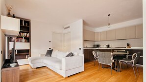 A modern living room with a white sofa, a dining area with a table and chairs, a kitchen with cabinets, and a bookshelf. - Hostmate - Milano - Ulpio Traiano (Milano)