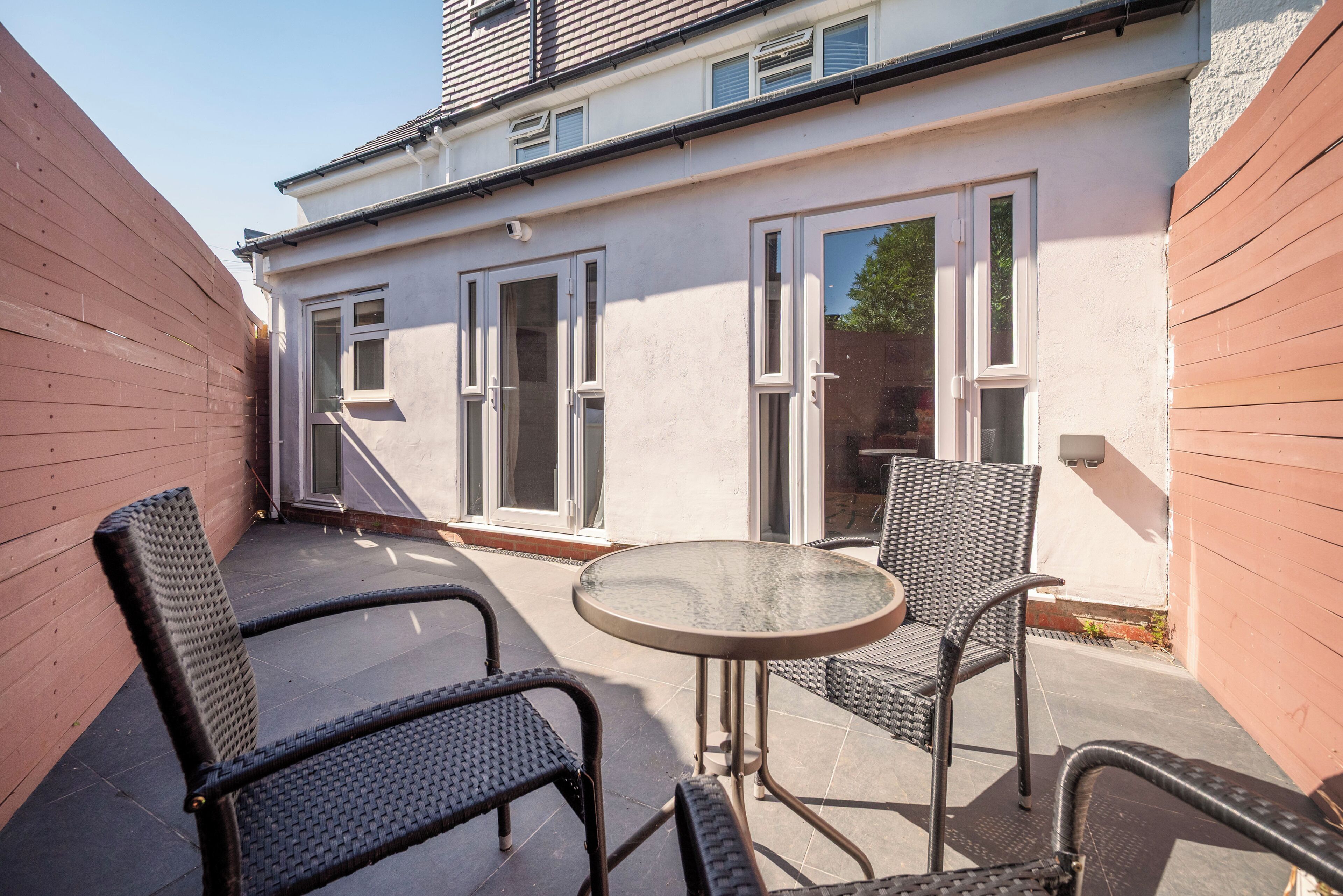 Une terrasse avec deux chaises et une table, un bâtiment blanc doté de grandes fenêtres et une clôture en bois.
