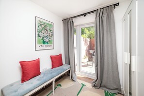 Une pièce avec un banc, des coussins rouges, une œuvre d’art encadrée, des rideaux gris et une vue à travers une porte vitrée.