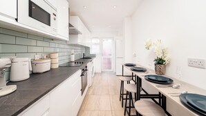 A modern kitchen with a microwave, stove, and various appliances. There is a dining area with a table and chairs. - Modern Luxury 4Bdrm 15 min to LDN centre | Business/Vacay | Parking onsite (London)