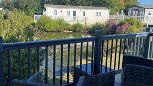 A deck with wicker furniture overlooking a body of water, a white building with a balcony, and a forested hill in the background. - DJD ,Country Style Luxury Lodge, Manor Park Sleeps 4 (Hunstanton)