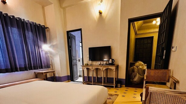 A hotel room with a large bed, a television on a wooden stand, a wicker chair, and a small table with a kettle. - SADRI HAVELI Boutique Heritage Hotel - Lake Pichola Udaipur (Udaipur)