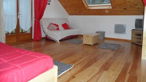 Um quarto aconchegante com teto de madeira, um sofá branco com almofadas vermelhas, uma cama com cobertor vermelho e uma televisão sobre um suporte de madeira.