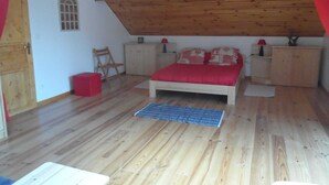 Quarto com teto inclinado, piso de madeira, cama com roupa de cama vermelha, duas mesas de cabeceira com luminárias, uma janela pequena e uma porta.