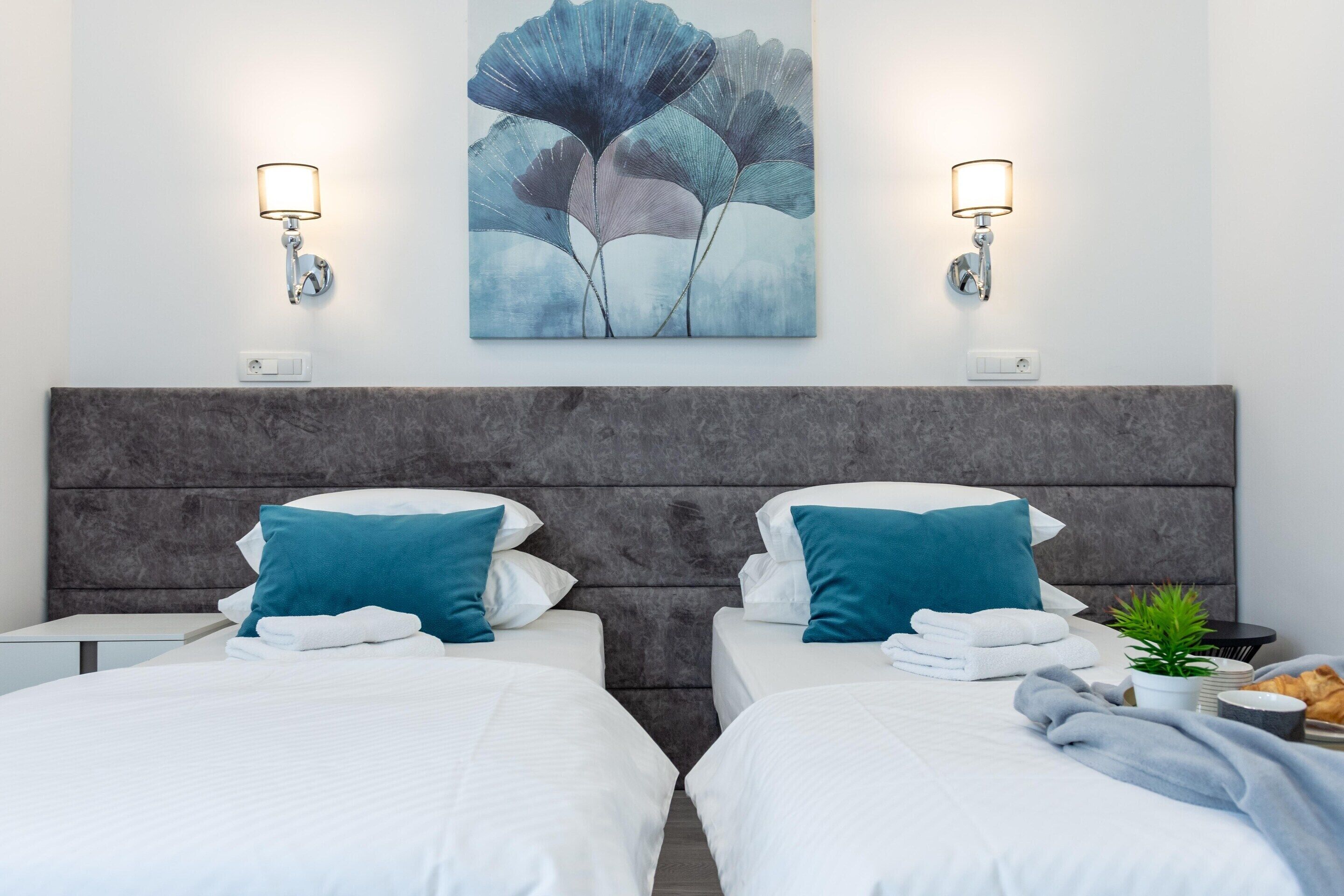 A hotel room with two beds, a grey headboard, wall-mounted lights, and a large abstract painting.