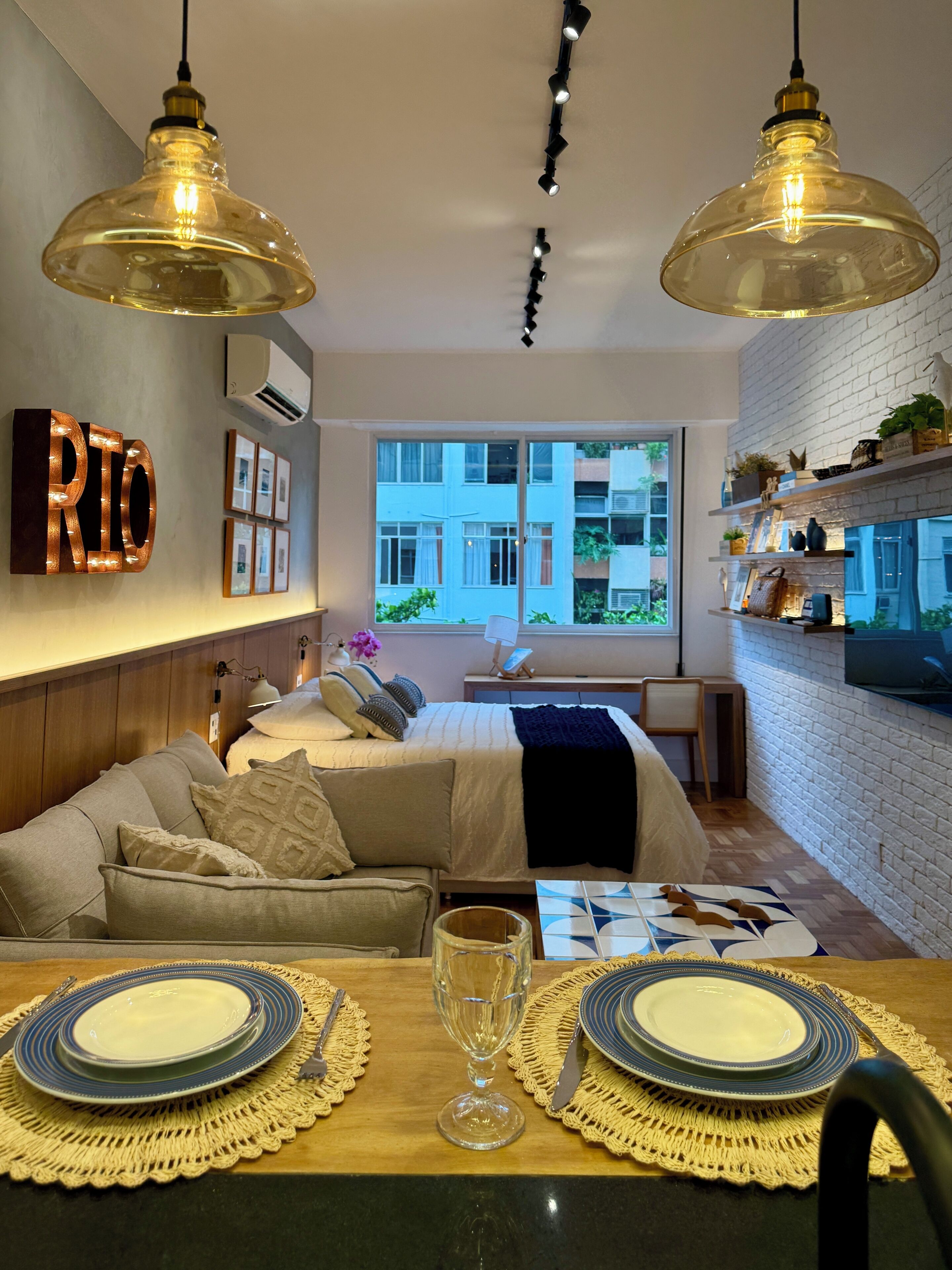 A modern bedroom with a sofa bed, a dining table with plates and a wine glass, and a large window with a view of buildings.
