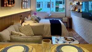 Quarto moderno com cama-sofá, mesa de jantar com pratos e taça de vinho, e uma grande janela com vista para edifícios.