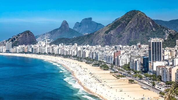 Paisagem costeira urbana com praia, edifícios altos e montanhas imponentes ao fundo.