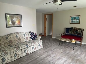 Un salon avec un canapé à motifs floraux, une chaise en bois avec un coussin rouge, un ventilateur de plafond et deux tableaux encadrés accrochés aux murs.