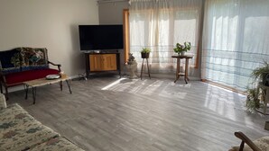 Una sala de estar con un sofá, televisor, ventilador de techo y un ventanal con cortinas traslúcidas.