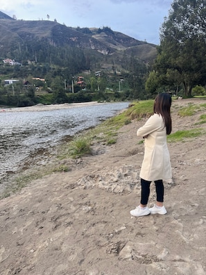 Una persona está de pie en una playa arenosa, con un río y colinas al fondo.