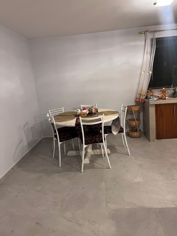 Un comedor con una mesa redonda, sillas blancas y una pequeña cocina con un lavamanos y armarios.