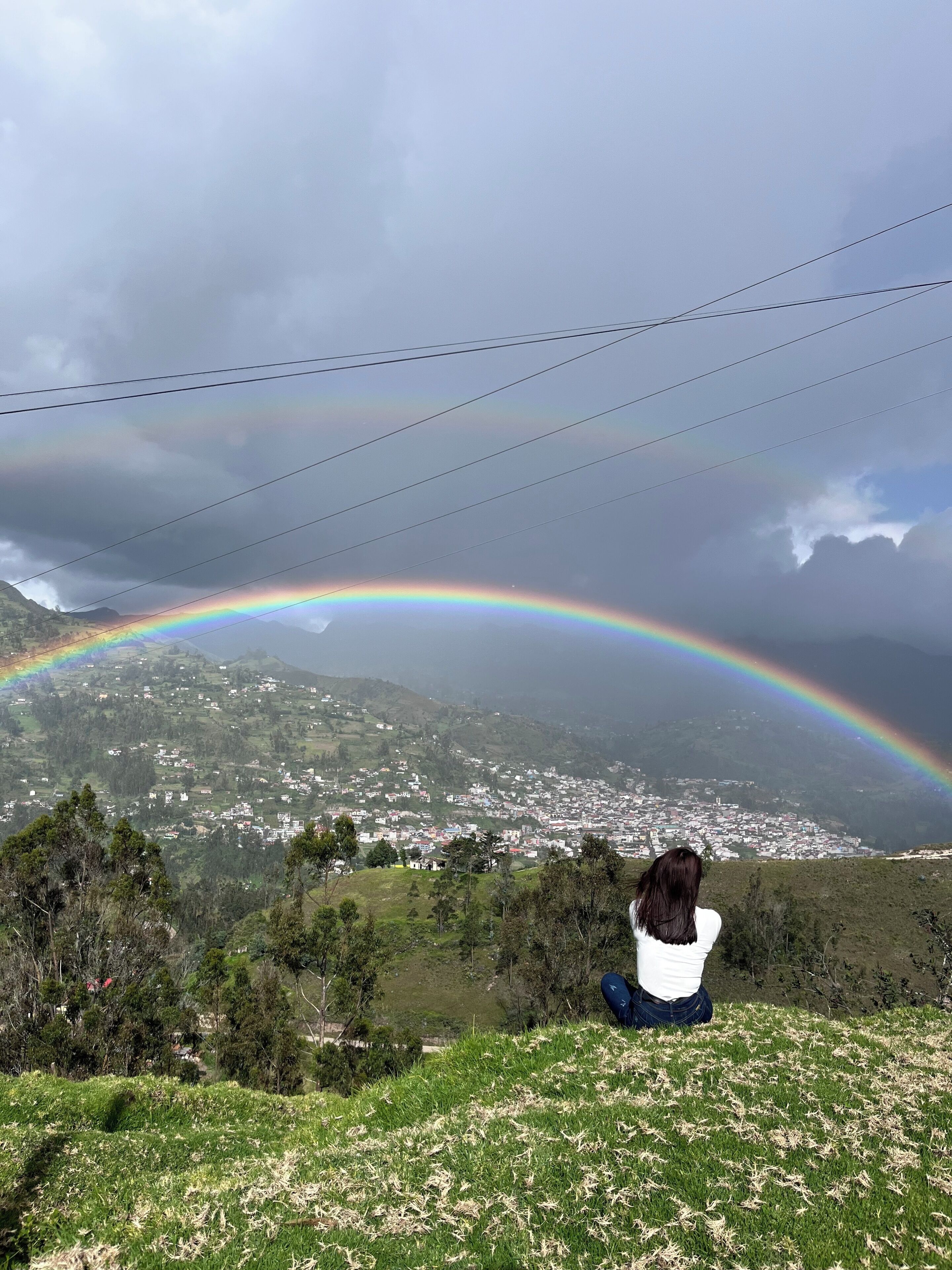 A person sitting on a grassy hill overlooking a town with a rainbow in the sky.