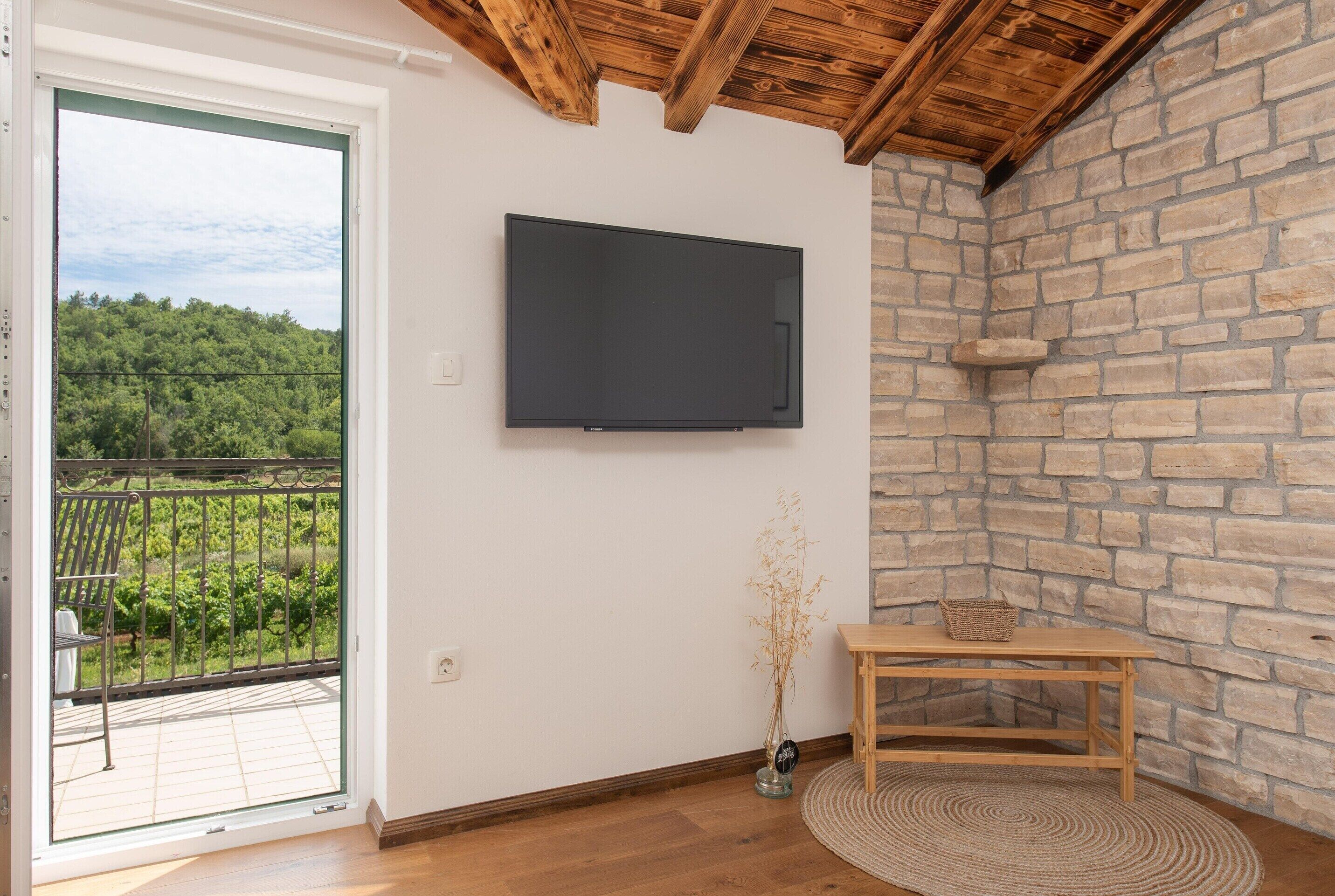 Une pièce avec un téléviseur fixé à un mur blanc, une table en bois avec un panier, un vase contenant des plantes séchées et une vue sur un paysage verdoyant par une porte vitrée.