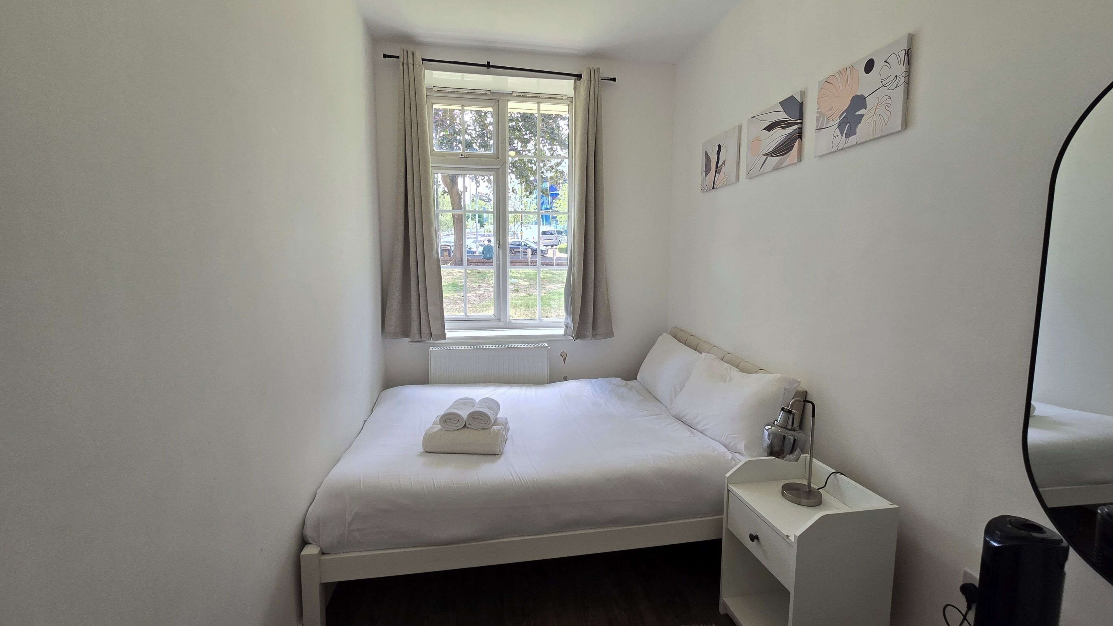 Een enkel bed met witte lakens, een nachtkastje met lamp en een raam met gordijnen.
