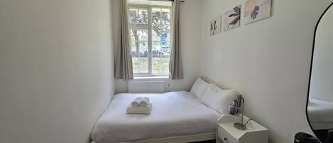 A single bed with white linens, a bedside table with a lamp, and a window with curtains.
