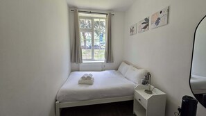 A single bed with white bedding, a bedside table with a lamp, and a window with curtains. - Free Parking + Modern 1BR Near Central London (Greater London)
