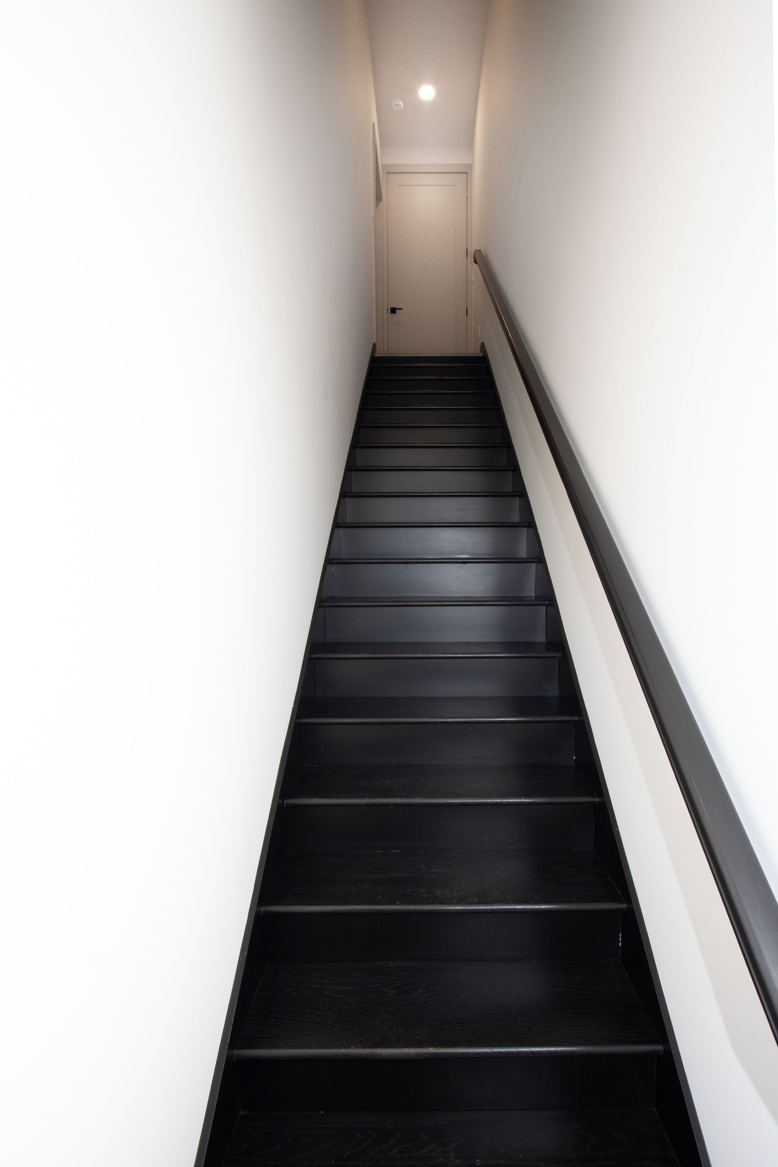 A narrow, straight staircase with black steps and white walls, leading to a door at the top.