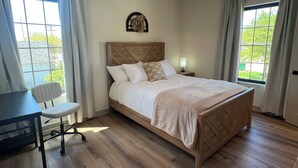 A neatly arranged bedroom with a wooden bed, a desk, and a chair, featuring large windows and a ceiling fan.