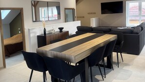 A modern living room with a dining area, featuring a wooden dining table, black chairs, a black sofa, and a TV mounted on the wall.