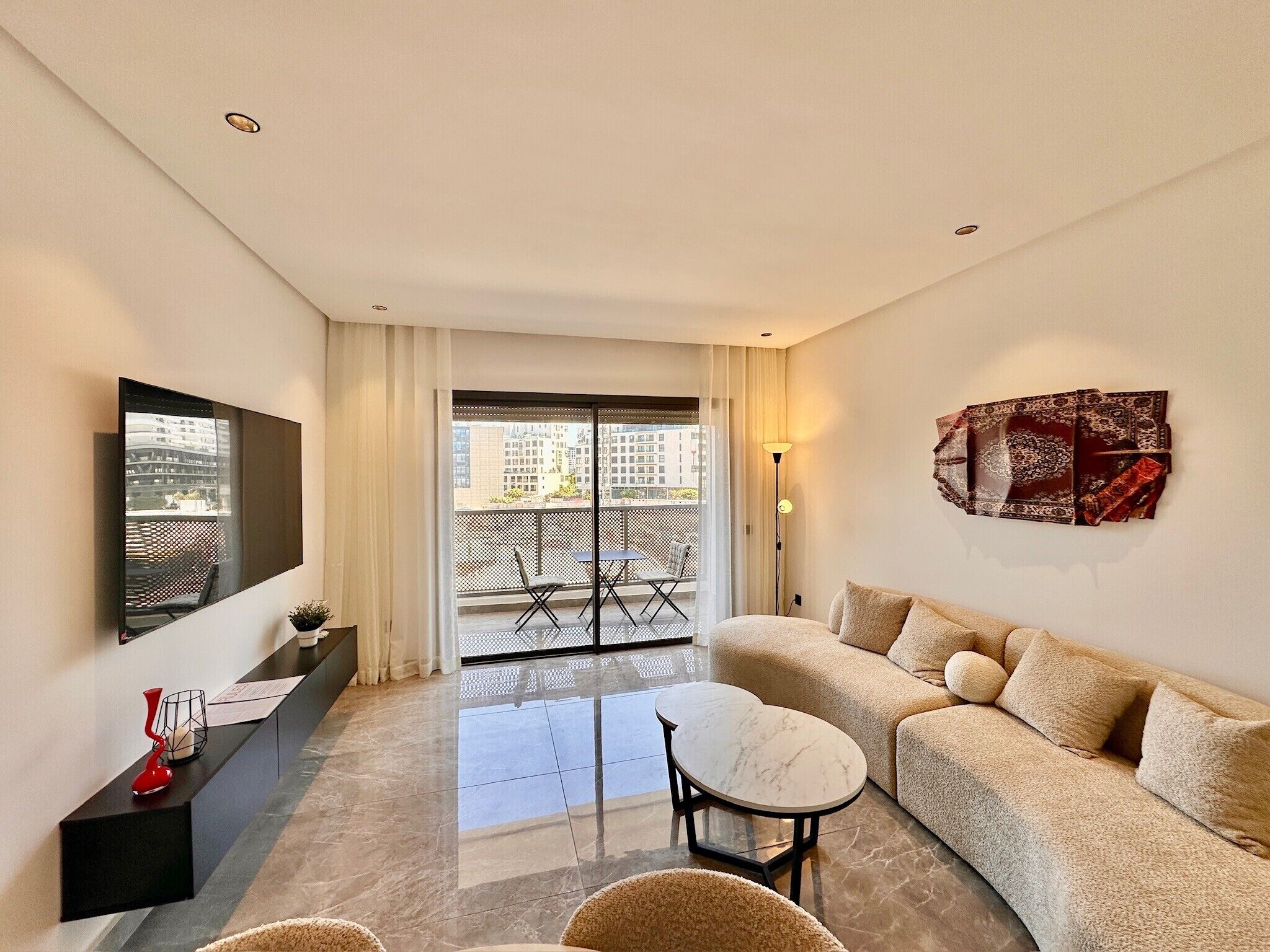 Un salon moderne avec un canapé, une petite table ronde, un téléviseur fixé au mur et un balcon offrant une vue sur les immebles.