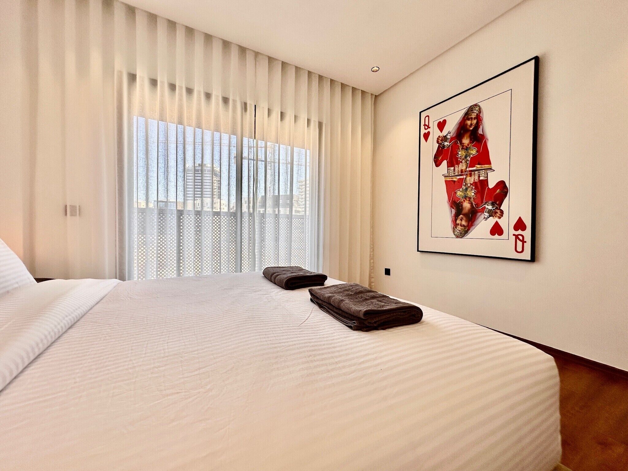 A hotel room with a large bed, a window with sheer curtains, and a framed playing card on the wall.