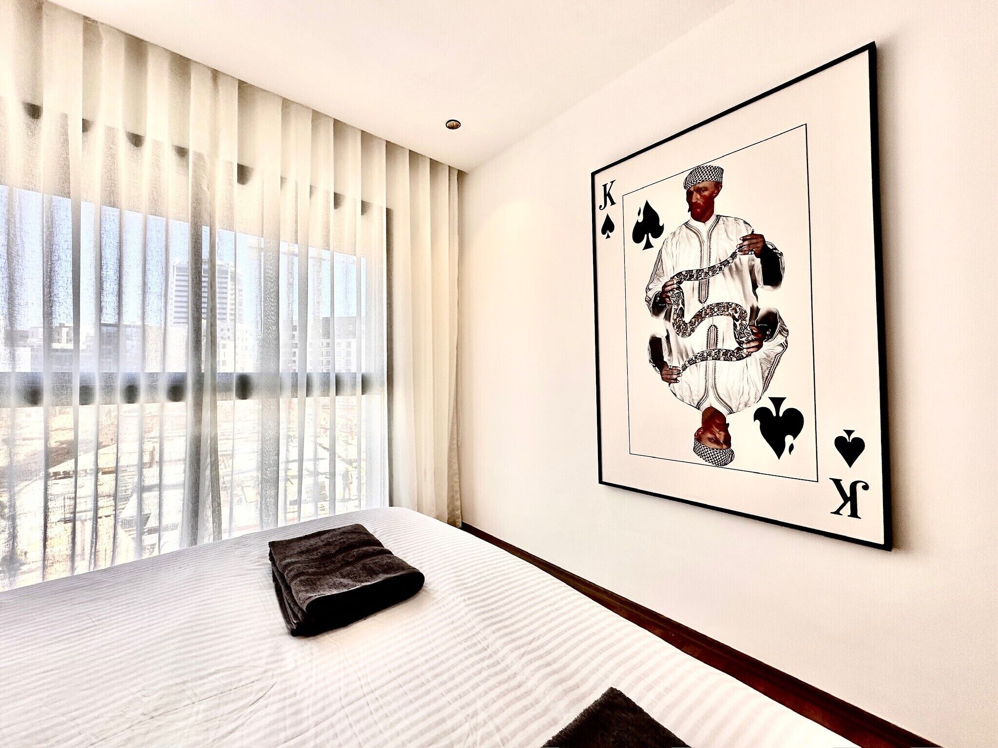 Une chambre d’hôtel avec un lit, une grande fenêtre avec des stores et une œuvre d’art représentant une carte à jouer, encadrée et accrochée au mur.