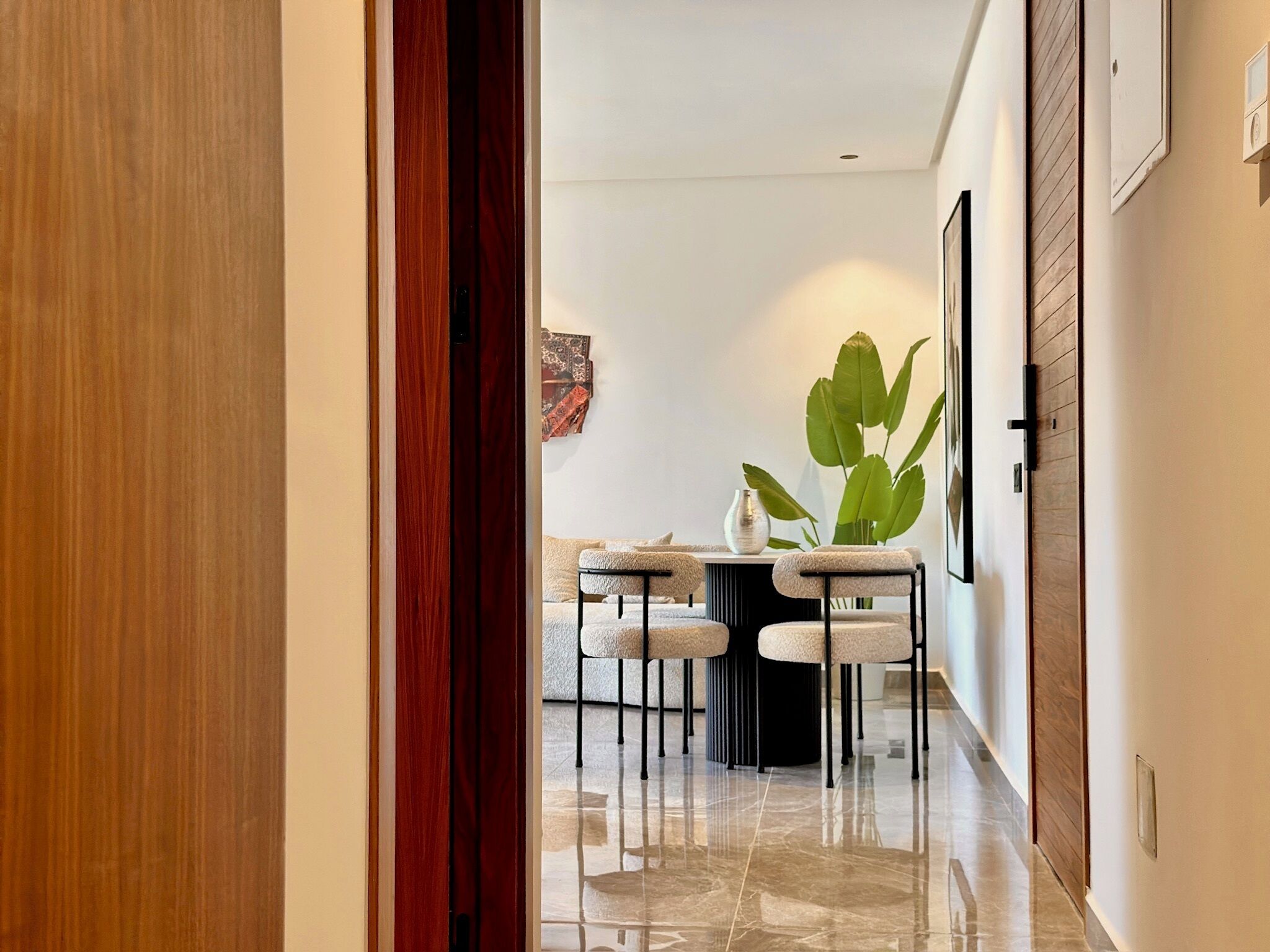 Un couloir mène à un espace repas comprenant une table et des chaises, une plante en pot et un luminaire fixé au mur.