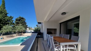 A modern outdoor patio with a pool, a dining area with white chairs and a wooden table, and a large window overlooking the pool. - Summer Color (Novalja)