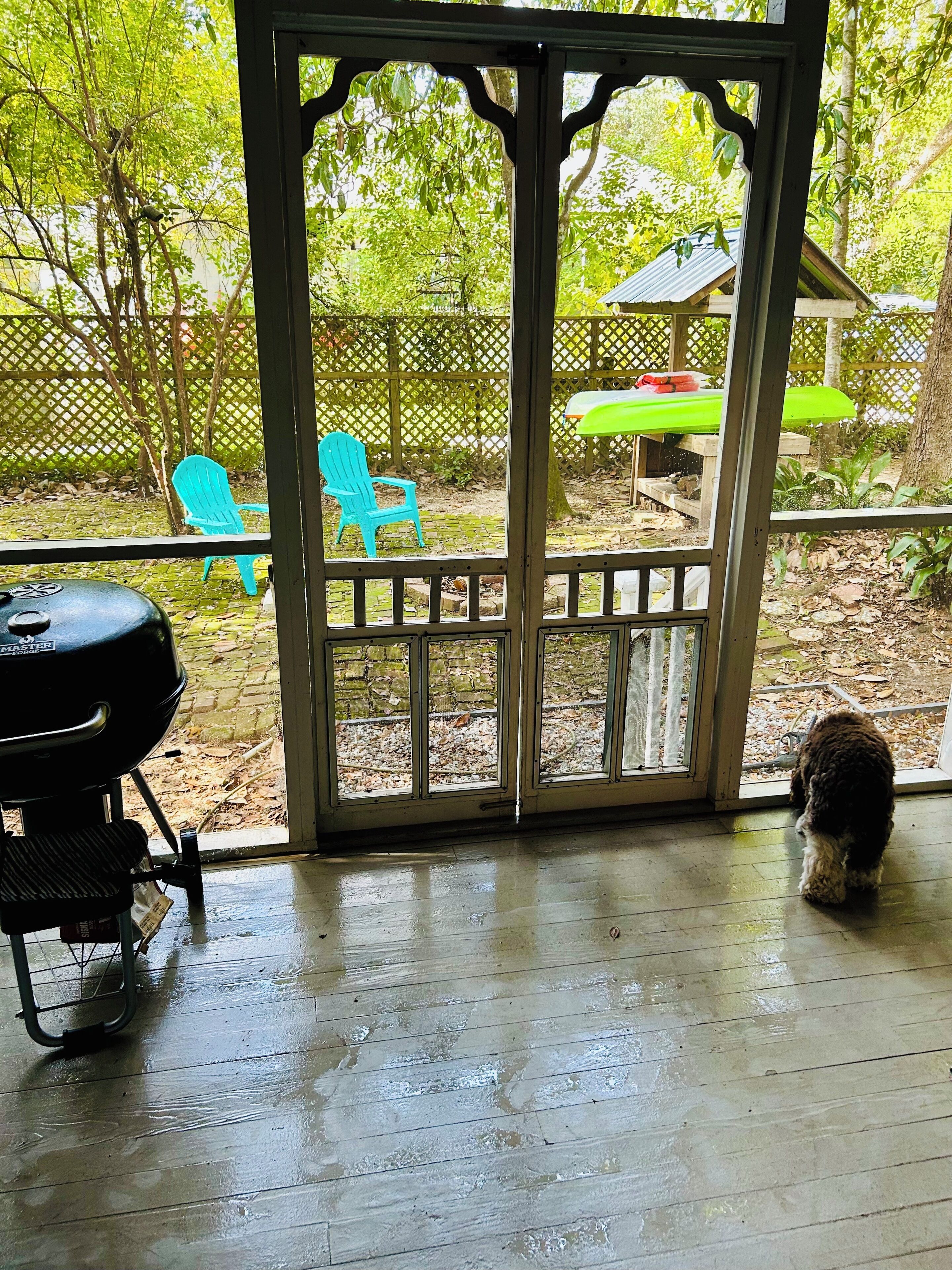A covered patio area with a grill, a wooden deck, and a view of a backyard with a chair and a table.