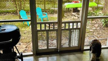 A covered patio area with a grill, a wooden deck, and a view of a backyard with a chair and a table.