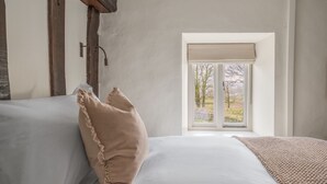 Quarto com uma janela grande, roupa de cama branca e vista para árvores e um muro de pedra.