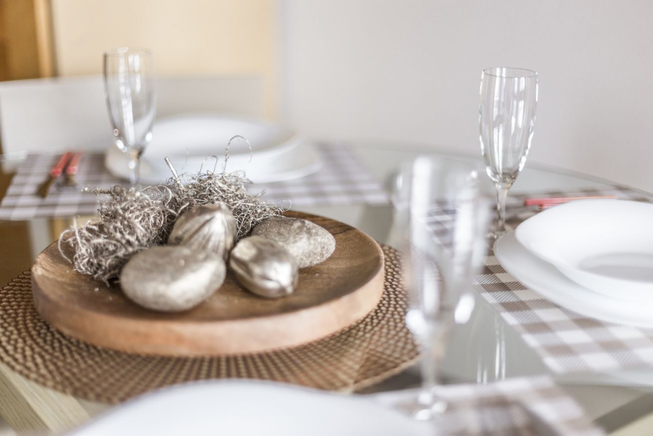 Una mesa de comedor dispuesta con platos blancos, cubiertos y un centro de mesa de madera con piedras y cuerda decorativa.