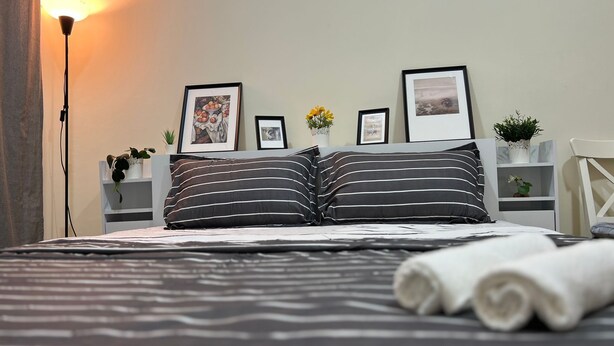 A neatly made bed with striped pillows, a bedside lamp, and framed pictures on the wall. - Beautiful Bedroom in AlBarsha Near Metro (Dubai)