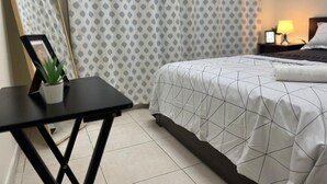 A hotel room with a bed, a small table with a plant, a nightstand with a lamp, and patterned curtains. - Beautiful Bedroom in AlBarsha Near Metro (Dubai)