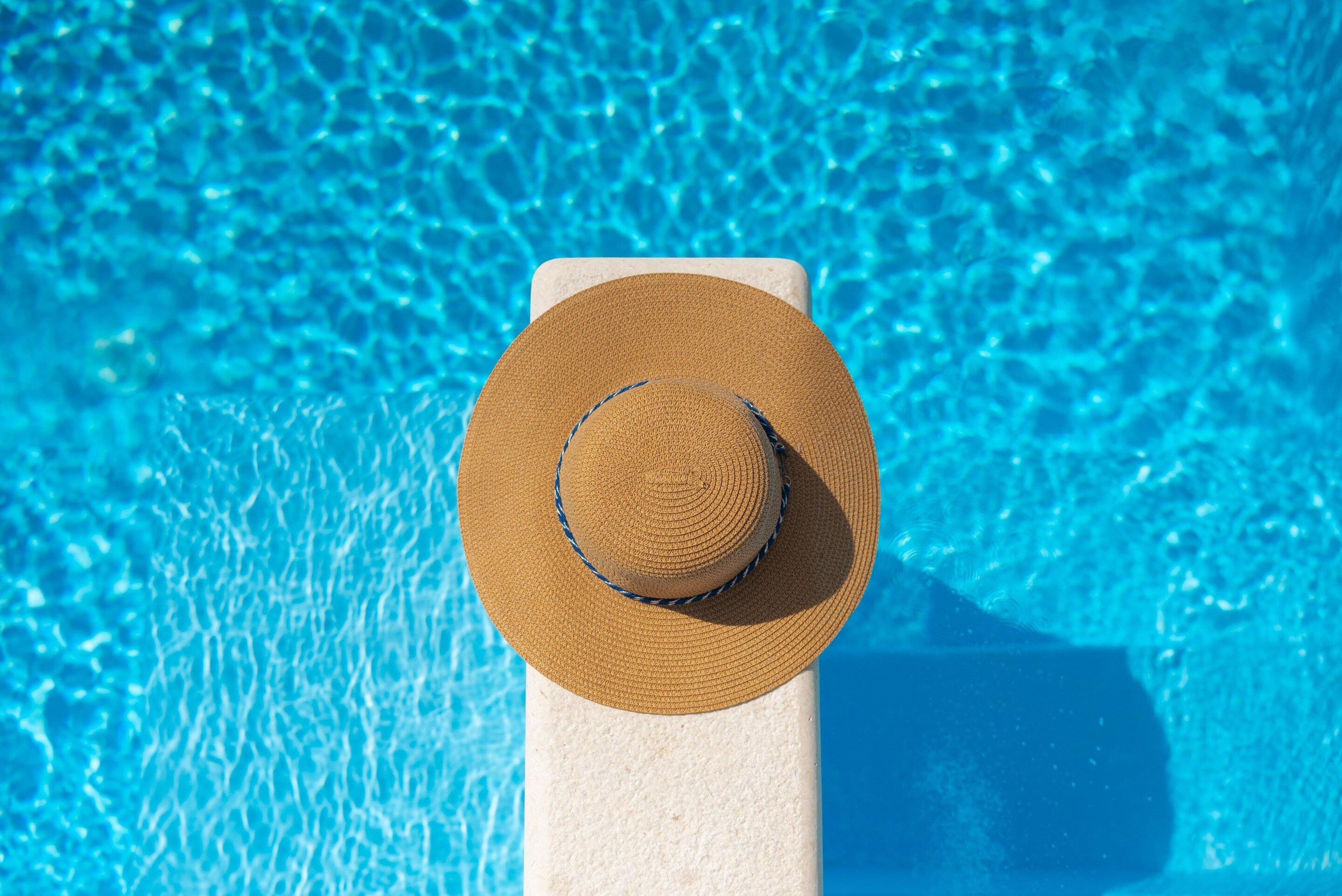 A straw hat on a white pedestal by a pool with clear blue water.