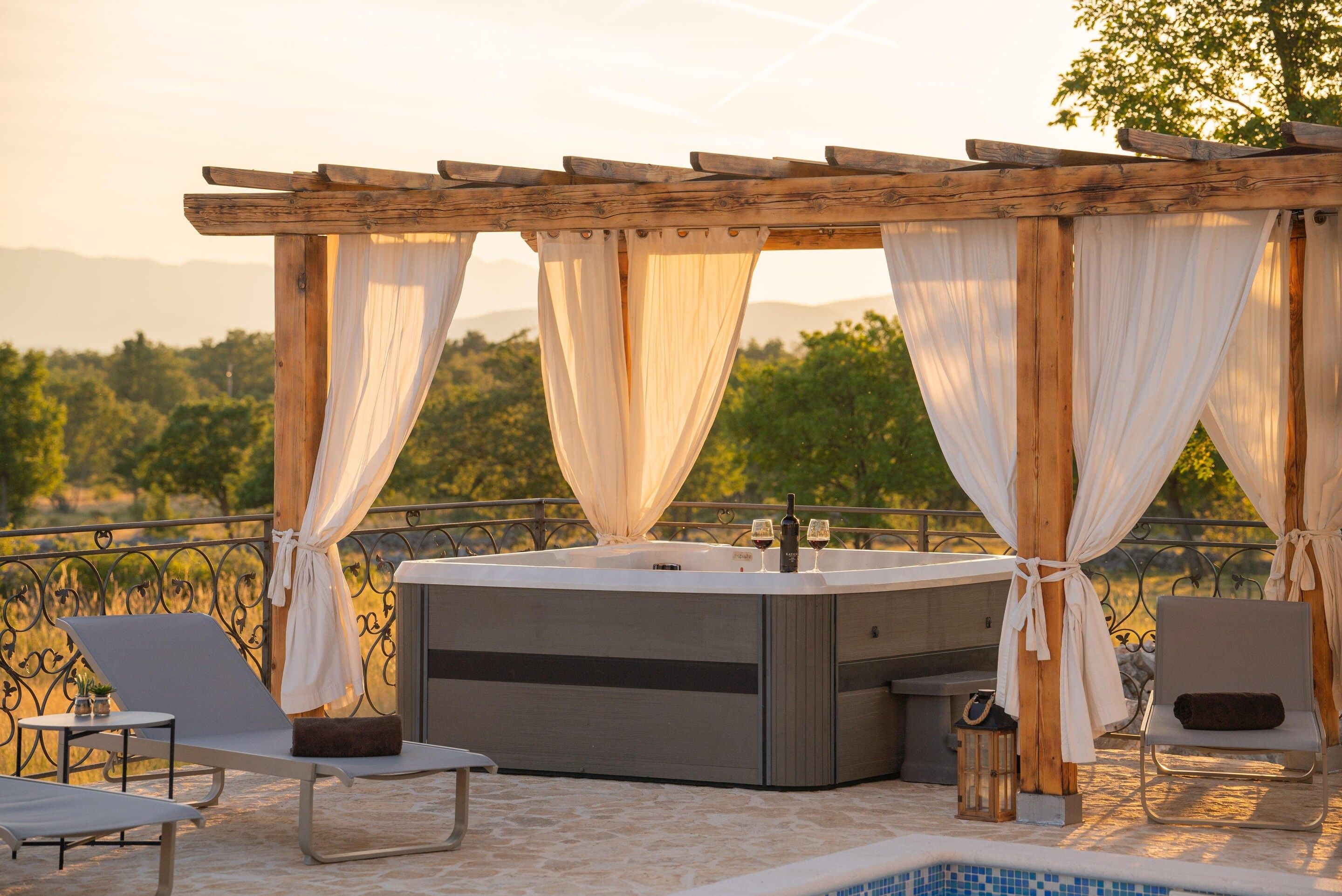 Ein Whirlpool mit einer Pergola und Vorhängen, zwei Liegestühle, ein kleiner Tisch mit einer Pflanze und Blick auf die Landschaft.